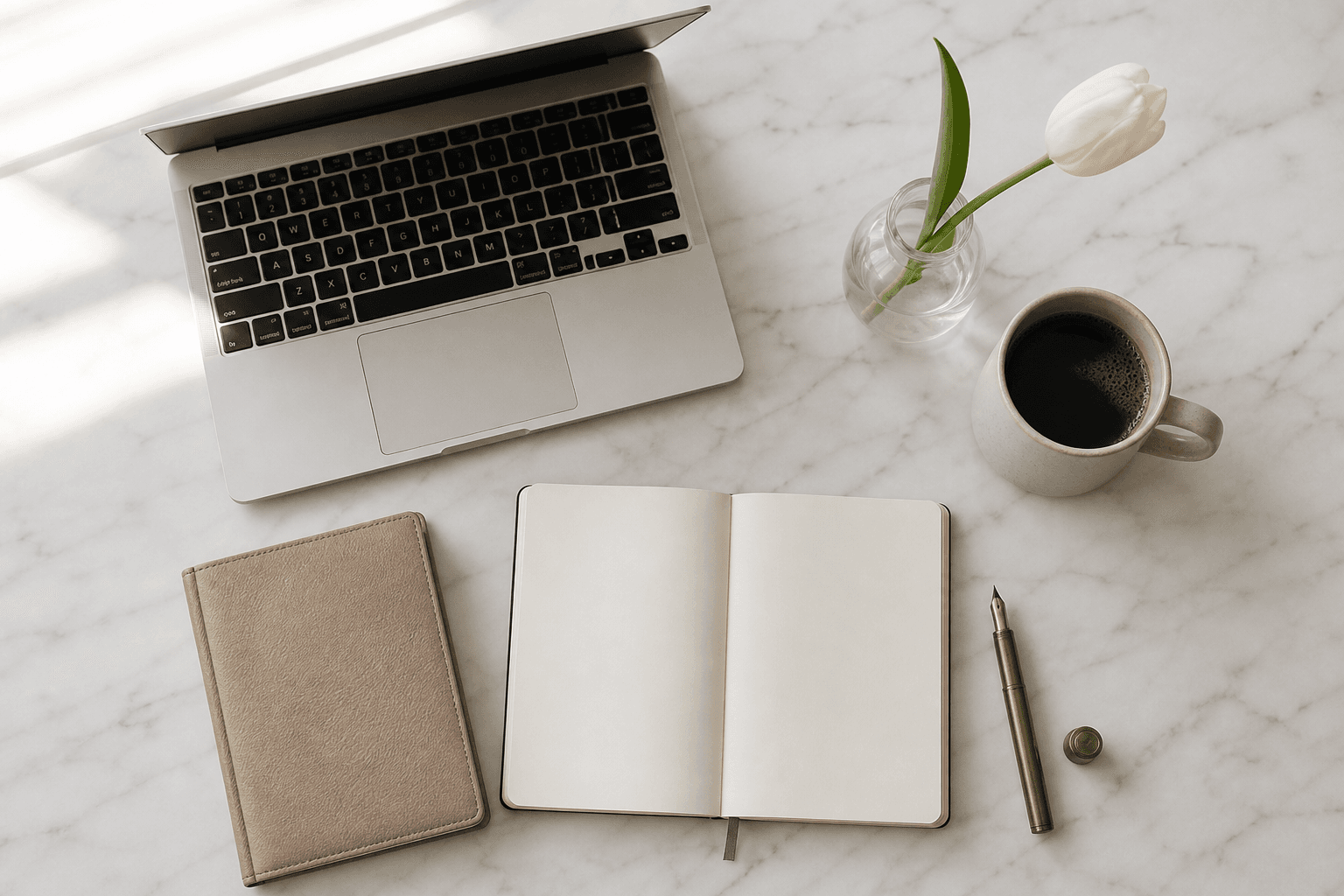 A pale marble desk shot from above with an open laptop, ceramic coffee cup, leather notebook, an open Moleskine, brass pen and a single white tulip in soft morning light.